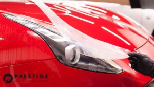 Close-up of a red sports car's headlight with a protective film being applied by a gloved hand, showcasing paint protection film installation. The Prestige Auto Appearance logo is visible in the corner, emphasizing the service provider