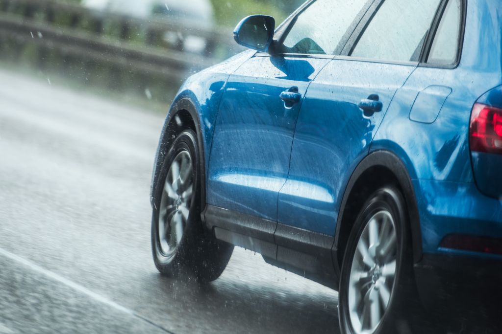 Blue SUV driving through heavy rain on a highway, with water spraying off the tires. The image illustrates challenging driving conditions in Pennsylvania, where road hazards like rain, road salt, and debris can damage vehicle paint.