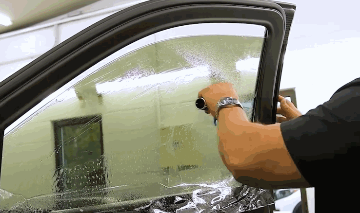 A professional applies a tinted film to the interior of a car window, using a squeegee to smooth out air bubbles. The process takes place in a clean automotive workshop.