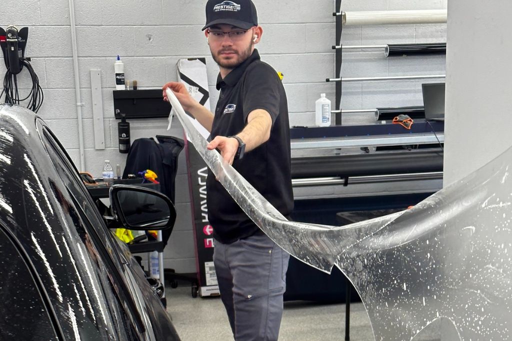 Technician at Prestige Auto Appearance holding a large sheet of clear paint protection film (PPF) while preparing to apply it to a black car. The image highlights professional PPF installation in a well-equipped workshop.