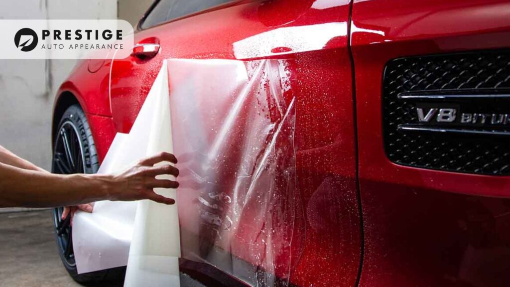 A close-up of a red luxury car with a "V8 Biturbo" badge, as a technician peels back a transparent paint protection film. The car's glossy surface reflects the surroundings. The "Prestige Auto Appearance" logo is visible in the corner.