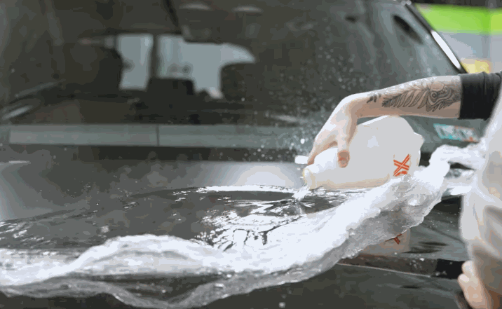 A technician with a tattooed arm pours soapy water over a car’s hood while applying a transparent paint protection film, ensuring smooth adhesion