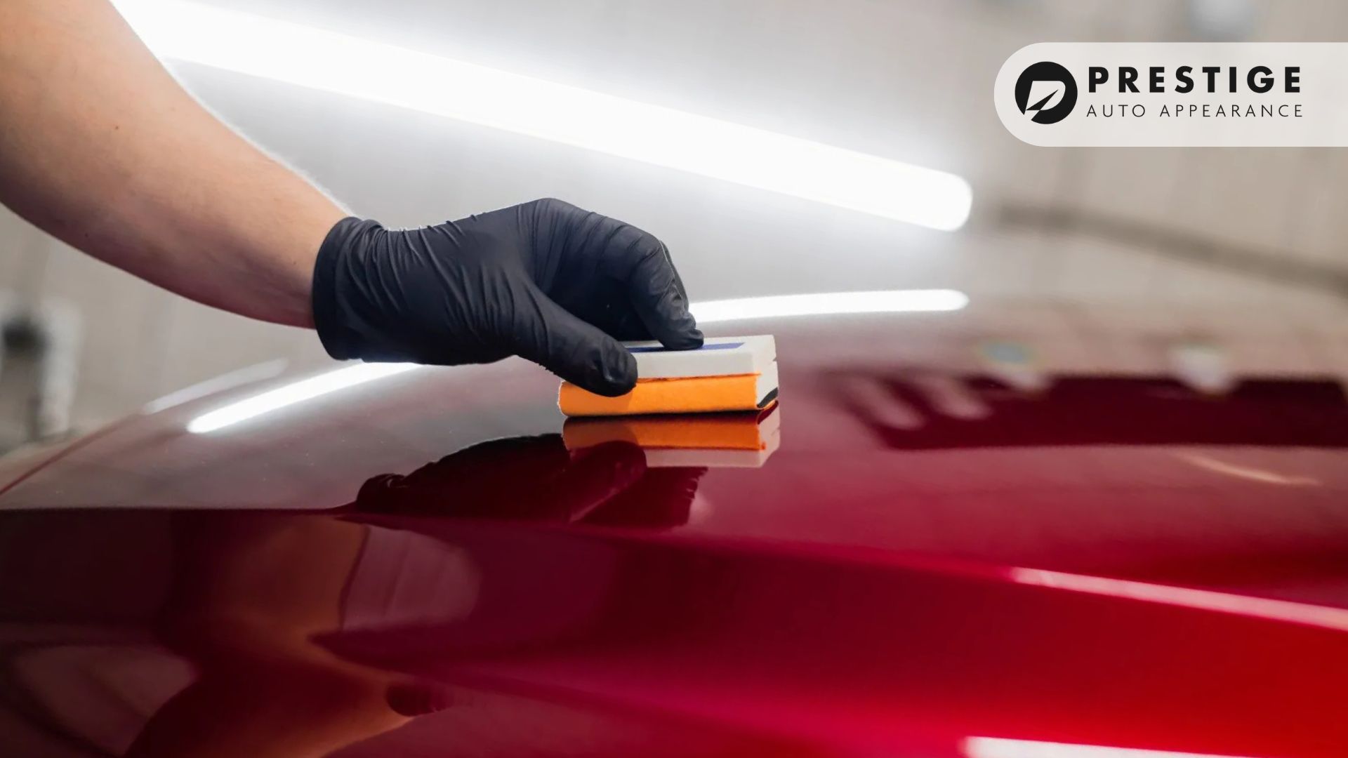 A technician wearing black gloves applies a ceramic coating to a glossy red car surface using an applicator pad